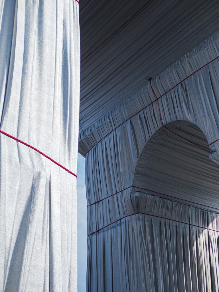 An abstract view of draped silver fabric with red accents, showcasing a structured archway and textured folds, set against a clear blue sky.