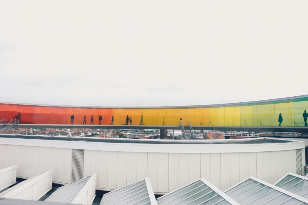 A colorful glass walkway curves above a cityscape, with silhouettes of people walking along it. The glass transitions from orange to yellow to green, creating a vibrant visual effect against a cloudy sky.