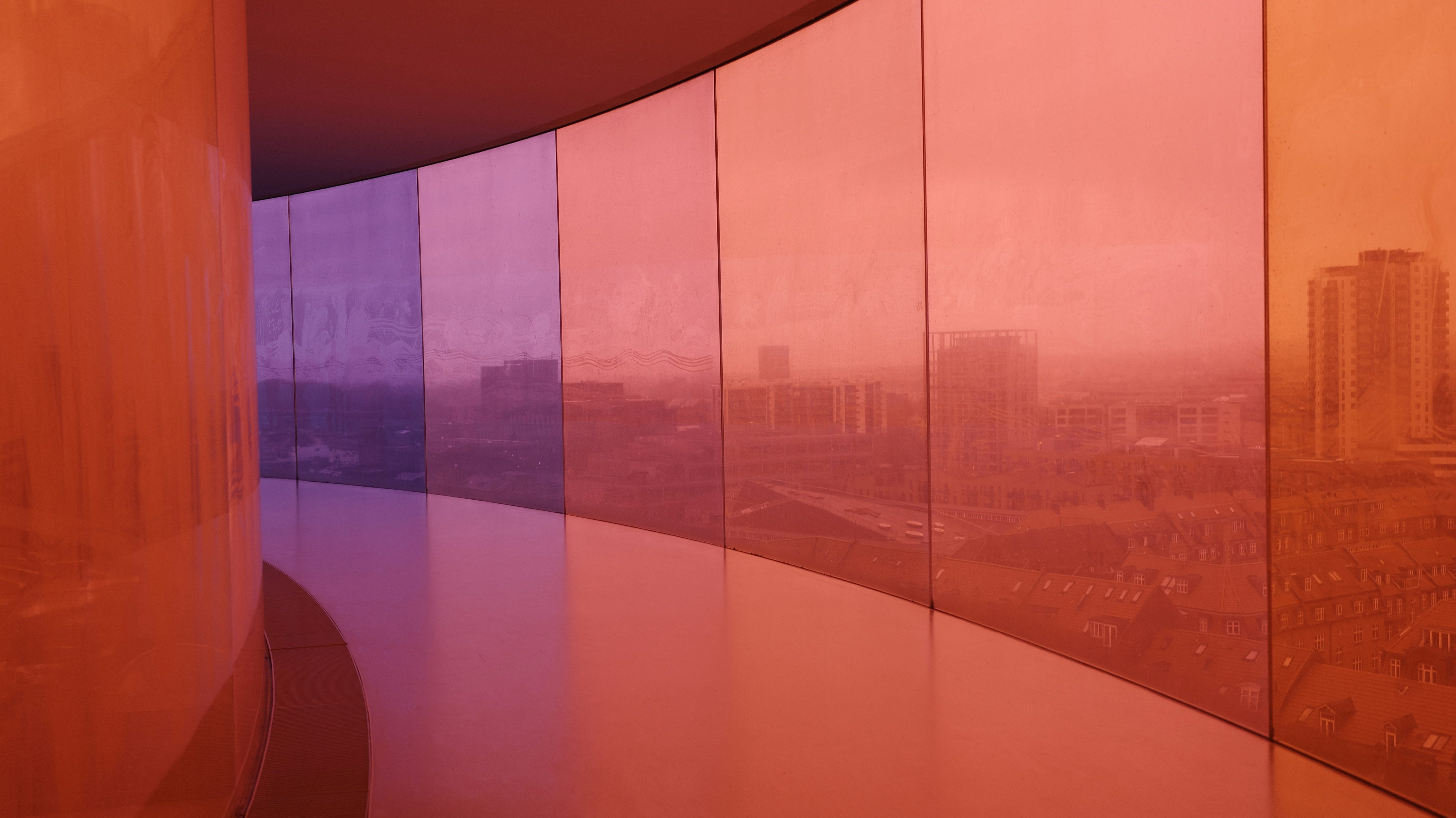 A curved interior space with large colored glass panels in shades of orange, purple, and pink, providing a view of the city skyline through a vibrant color filter.